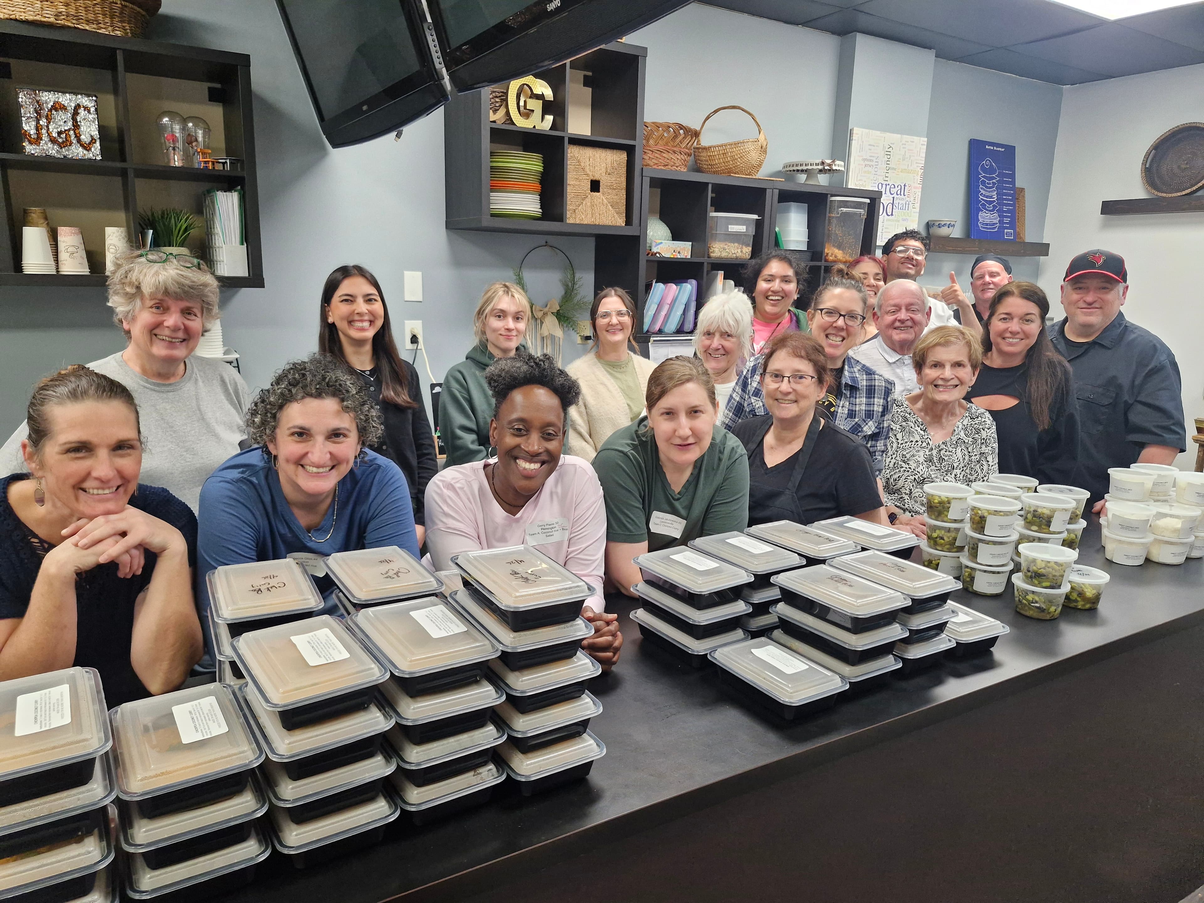 A diverse group of smiling people posing behind a counter stacked with prepared meal containers.