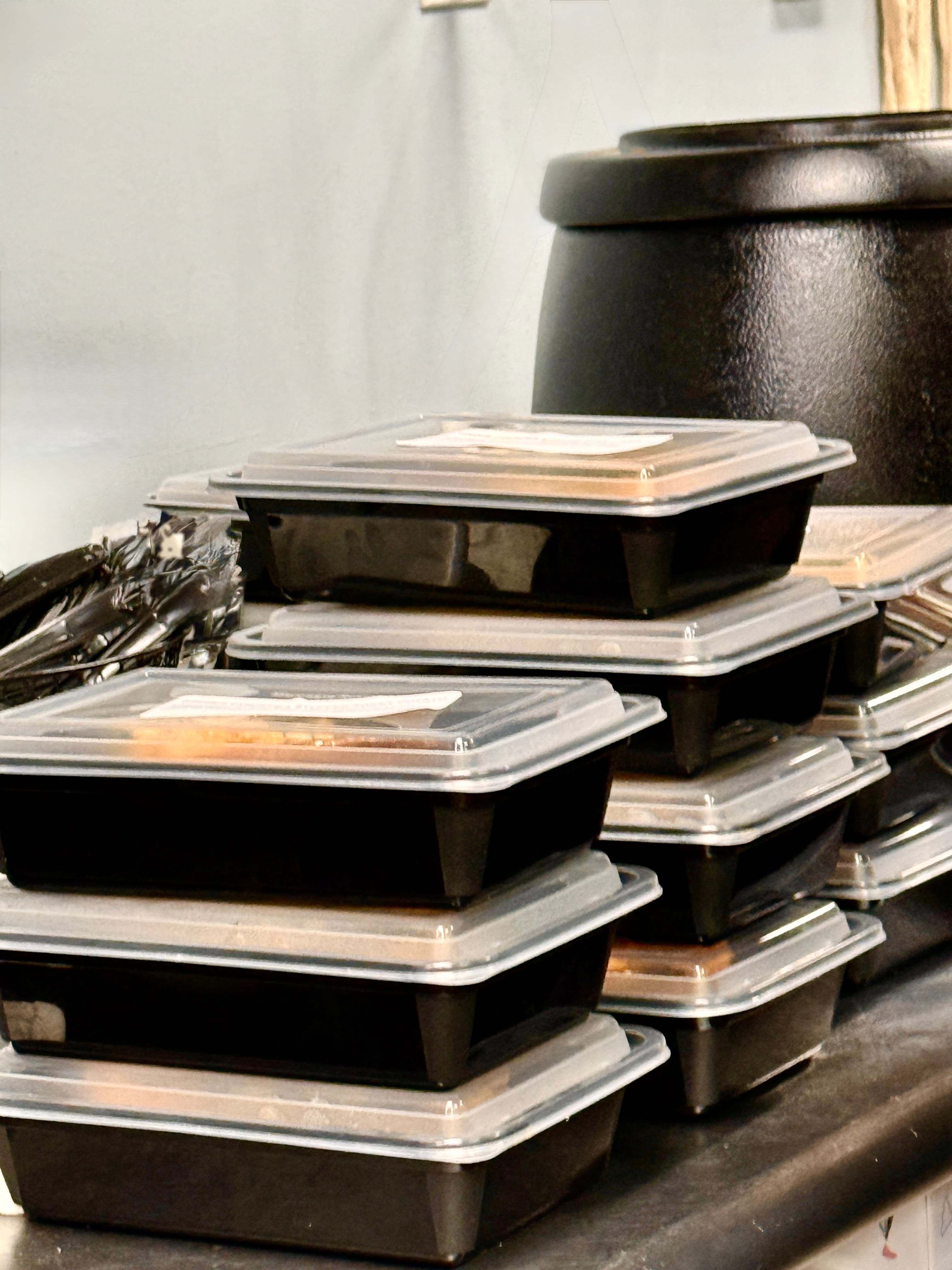 Stacks of black plastic food containers with clear lids beside a large black pot.