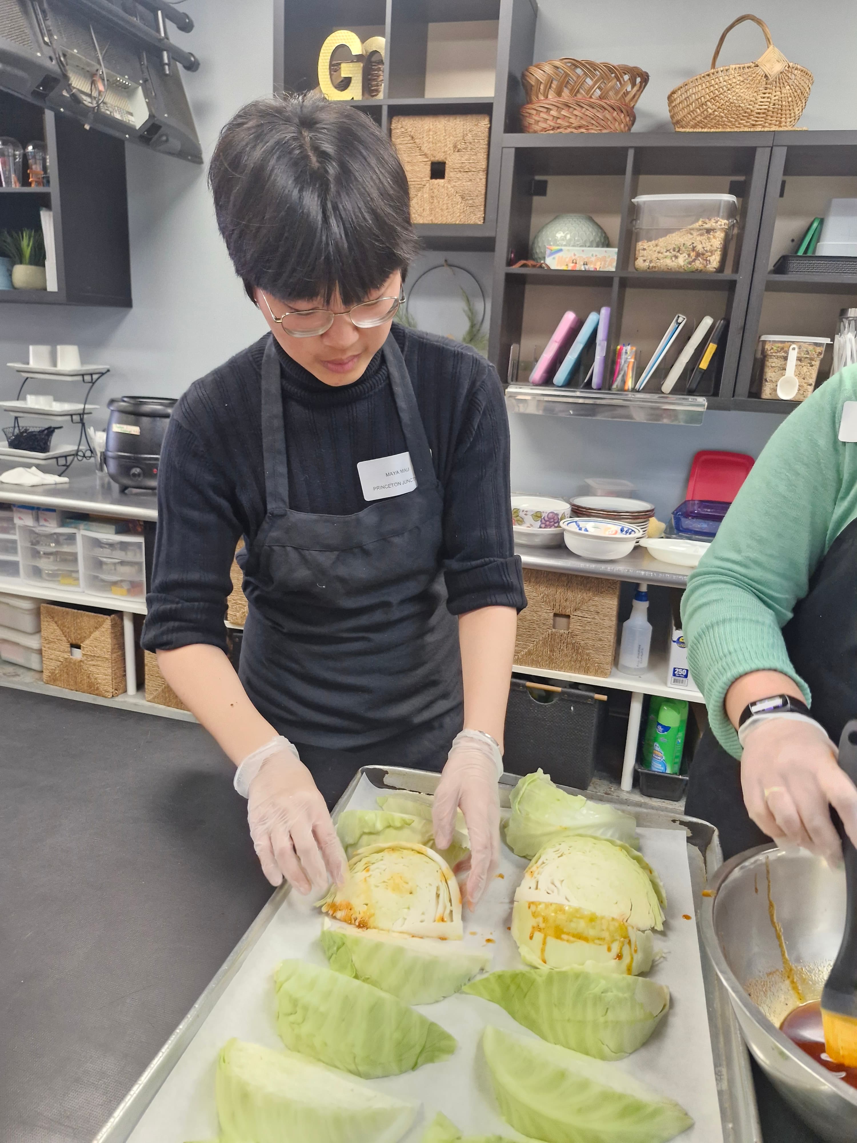 Person in black apron and gloves prepares cabbage wedges with sauce on a baking sheet.