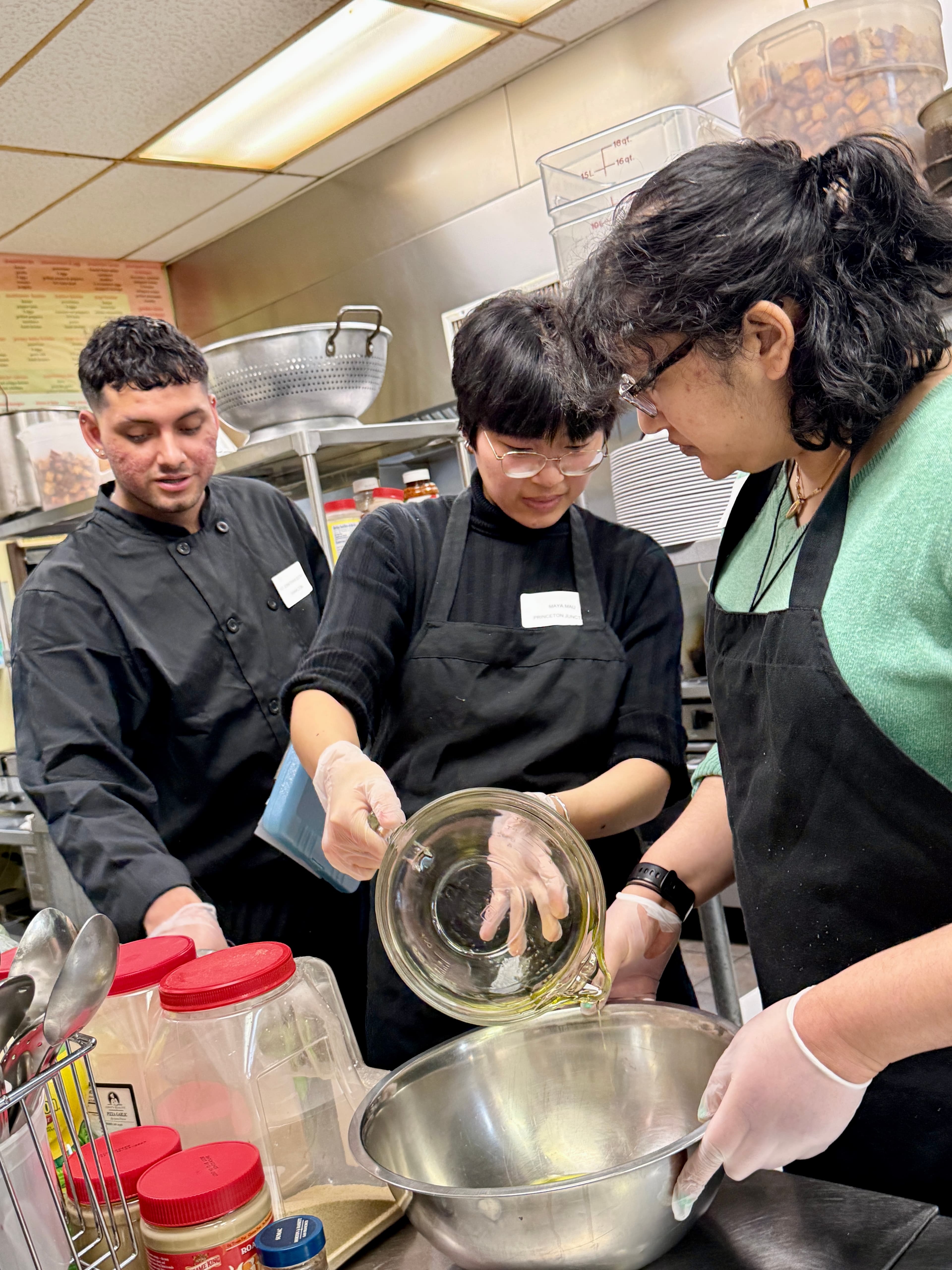 Three kitchen workers in aprons and gloves pour liquid into a large metal bowl.
